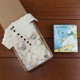 Luxury Aran & Bedtime Book Gift. A gift box containing a cream-colored Aran cardigan with buttons, a matching hat, and mittens sits on a wooden surface. Next to the box is the children's book "The Children of Lir," featuring an illustration of a swan on the cover. Luxury Aran & Bedtime Book Gift. A gift box containing a cream-colored Aran cardigan with buttons, a matching hat, and mittens sits on a wooden surface. Next to the box is the children's book "The Children of Lir," featuring an illustration of a swan on the cover.